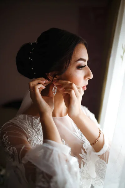 Stylish happy bride is getting ready in the morning. Happy newlywed woman in bedroom. Beautiful woman with professional make up and hair style. Morning of the bride. Wedding day. Fashion bride.