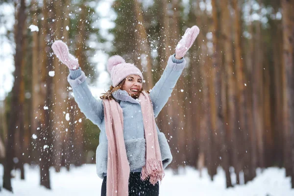 Cute young woman  is playing with snow  in the forest. The girl is dressed in a blue coat, in knitted hat, scarf and mittens. Winter lifestyle happiness emotions nature. Christmas, New year.