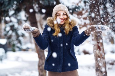 Güzel kız kış ormanında karla oynuyor. Mavi ceketli, örgü şapkalı ve eldivenli gülümseyen kız, kar ellere düşerken eğleniyor. Kış parkında moda genç bayan. Noel. 