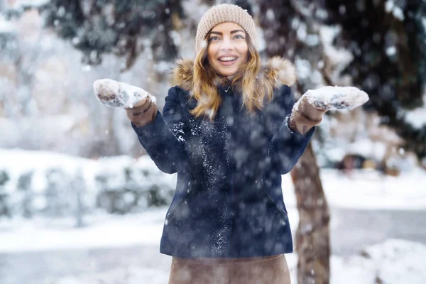 美丽的女孩在冬天的森林里与雪玩耍 穿着蓝色夹克 头戴针织帽子 头戴手套 手里拿着雪开心地笑着的女孩 在冬季公园里的时髦的年轻女子 圣诞节 — 图库照片