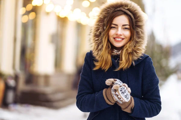 穿着蓝色夹克 头戴针织帽子 头戴手套的漂亮姑娘在街上摆姿势 加兰德灯 喜欢雪 对着相机微笑 快乐的新年心情 真实的情感 冬季时尚 — 图库照片