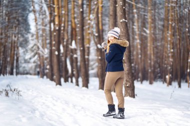 Uzun saçlı, mavi ceketli, örgü şapkalı ve eldivenli güneşli kış gününde poz veren güzel bir kızın portresi. Kış ormanlarında moda genç kadın. Noel, kış tatili konsepti. 