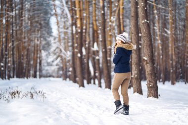 Uzun saçlı, mavi ceketli, örgü şapkalı ve eldivenli güneşli kış gününde poz veren güzel bir kızın portresi. Kış ormanlarında moda genç kadın. Noel, kış tatili konsepti. 