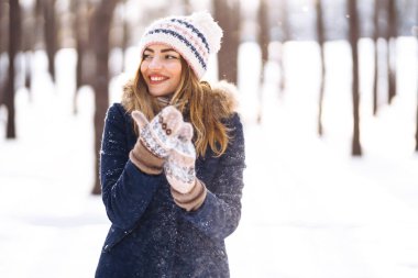 Uzun saçlı, mavi ceketli, örgü şapkalı ve eldivenli güneşli kış gününde poz veren güzel bir kızın portresi. Kış ormanlarında moda genç kadın. Noel, kış tatili konsepti. 