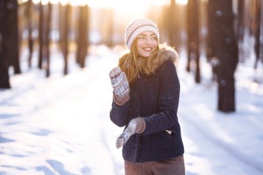 Mavi ceketli, örgü şapkalı ve eldivenli güzel bir kızın portresi kış ormanında gün batımında poz veriyor. Kameraya gülümsüyor, neşeli yeni yıl havası, gerçek duygular. Kış modası. Noel. 