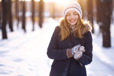 Mavi ceketli, örgü şapkalı ve eldivenli güzel bir kızın portresi kış ormanında gün batımında poz veriyor. Kameraya gülümsüyor, neşeli yeni yıl havası, gerçek duygular. Kış modası. Noel. 
