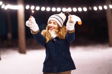 Kış ormanında elinde maytap tutan neşeli genç bir kadın. Maytapla poz veren örgü şapkalı mutlu şirin kız. Bengal ışığıyla yeni yıl arifesi. Kadın mutluluğu ve havai fişek oyunu.