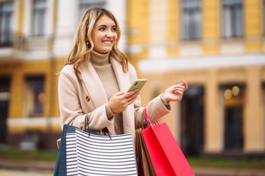 Telefonu olan güzel bir kadın şehir caddesinde alışveriş yapıyor. Sokakta renkli alışveriş torbalarıyla alışveriş yapan mutlu bir kız. Tüketim, satın alma, alışveriş, yaşam tarzı konsepti.