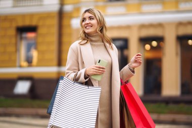 Telefonu olan güzel bir kadın şehir caddesinde alışveriş yapıyor. Sokakta renkli alışveriş torbalarıyla alışveriş yapan mutlu bir kız. Tüketim, satın alma, alışveriş, yaşam tarzı konsepti.