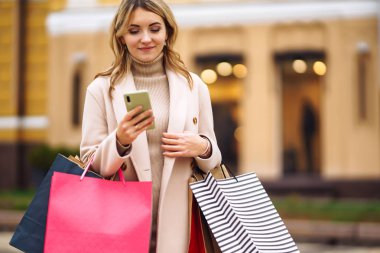 Telefonu olan güzel bir kadın şehir caddesinde alışveriş yapıyor. Sokakta renkli alışveriş torbalarıyla alışveriş yapan mutlu bir kız. Tüketim, satın alma, alışveriş, yaşam tarzı konsepti.