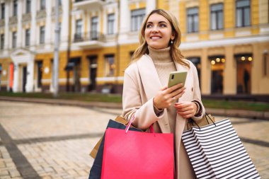 Telefonu olan güzel bir kadın şehir caddesinde alışveriş yapıyor. Sokakta renkli alışveriş torbalarıyla alışveriş yapan mutlu bir kız. Tüketim, satın alma, alışveriş, yaşam tarzı konsepti.