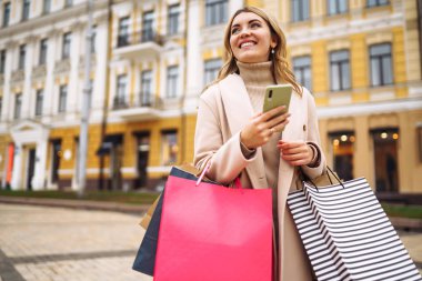 Telefonu olan güzel bir kadın şehir caddesinde alışveriş yapıyor. Sokakta renkli alışveriş torbalarıyla alışveriş yapan mutlu bir kız. Tüketim, satın alma, alışveriş, yaşam tarzı konsepti.