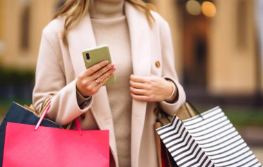 Elinde telefon ve alışveriş torbaları olan güzel bir kadının elleri. Sokakta renkli alışveriş torbalarıyla alışveriş yapan mutlu bir kız. Tüketim, satın alma, alışveriş, yaşam tarzı konsepti.