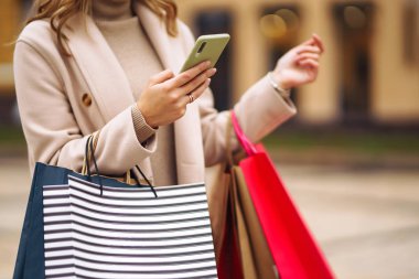 Elinde telefon ve alışveriş torbaları olan güzel bir kadının elleri. Sokakta renkli alışveriş torbalarıyla alışveriş yapan mutlu bir kız. Tüketim, satın alma, alışveriş, yaşam tarzı konsepti.