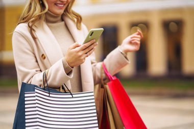 Elinde telefon ve alışveriş torbaları olan güzel bir kadının elleri. Sokakta renkli alışveriş torbalarıyla alışveriş yapan mutlu bir kız. Tüketim, satın alma, alışveriş, yaşam tarzı konsepti.