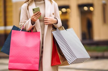 Elinde telefon ve alışveriş torbaları olan güzel bir kadının elleri. Sokakta renkli alışveriş torbalarıyla alışveriş yapan mutlu bir kız. Tüketim, satın alma, alışveriş, yaşam tarzı konsepti.