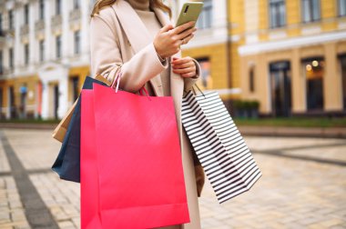 Elinde telefon ve alışveriş torbaları olan güzel bir kadının elleri. Sokakta renkli alışveriş torbalarıyla alışveriş yapan mutlu bir kız. Tüketim, satın alma, alışveriş, yaşam tarzı konsepti.