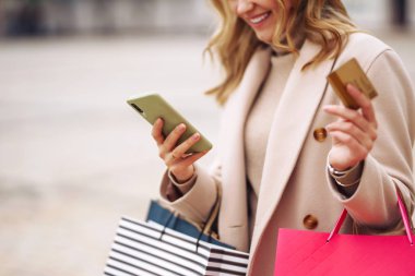 Telefonu ve kredi kartı olan güzel bir kadının elleri. Şehir caddesinde alışveriş torbaları tutan genç bir kadın. İnternetten alışveriş konsepti. Tüketim, satın alma, alışveriş, yaşam tarzı konsepti.