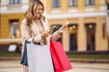 Telefonu ve kredi kartı olan güzel bir kadın. Şehir caddesinde elinde alışveriş torbaları tutan açık renk ceketli sarışın bir kız. İnternetten alışveriş konsepti. Tüketim, satın alma, alışveriş, yaşam tarzı konsepti.