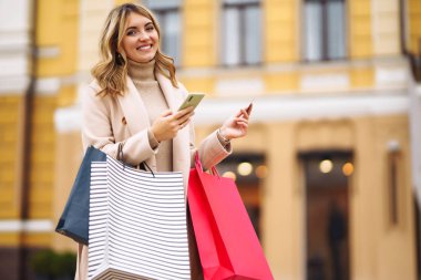 Telefonu ve kredi kartı olan güzel bir kadın. Şehir caddesinde elinde alışveriş torbaları tutan açık renk ceketli sarışın bir kız. İnternetten alışveriş konsepti. Tüketim, satın alma, alışveriş, yaşam tarzı konsepti.