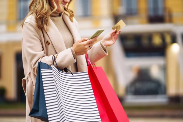 Hands of beautiful woman with telephone and a credit card. Young woman holding shopping bags on the city street. Online shopping concept. Consumerism, purchases, shopping, lifestyle concept.