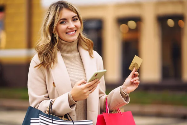 Telefonu ve kredi kartı olan güzel bir kadın. Şehir caddesinde elinde alışveriş torbaları tutan açık renk ceketli sarışın bir kız. İnternetten alışveriş konsepti. Tüketim, satın alma, alışveriş, yaşam tarzı konsepti.
