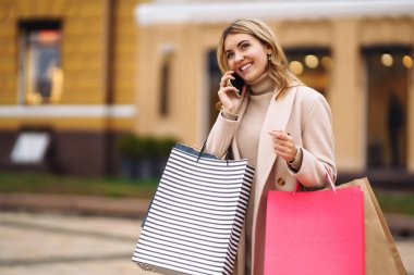 Alışveriş torbaları tutan ve telefonda konuşan genç bir kadın. Açık renk ceketli sarışın kız gülümsüyor ve renkli alışveriş torbalarını tutuyor. Tüketim, satın alma, alışveriş, satış, yaşam tarzı konsepti.