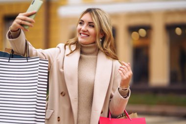 Alışveriş torbalı güzel bir kız selfie çekiyor. Şehirde alışveriş torbaları tutan genç bir kadın telefonda selfie çekiyor. Tüketim, satın alma, alışveriş, yaşam tarzı konsepti.