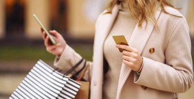 Telefonu ve kredi kartı olan güzel bir kadının elleri. Şehir caddesinde alışveriş torbaları tutan genç bir kadın. İnternetten alışveriş konsepti. Tüketim, satın alma, alışveriş, yaşam tarzı konsepti.
