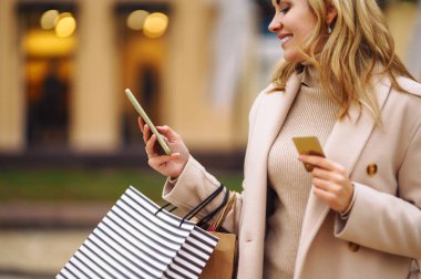 Telefonu ve kredi kartı olan güzel bir kadının elleri. Şehir caddesinde alışveriş torbaları tutan genç bir kadın. İnternetten alışveriş konsepti. Tüketim, satın alma, alışveriş, yaşam tarzı konsepti.