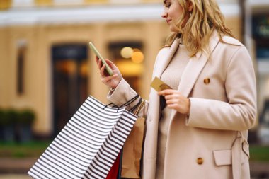 Telefonu ve kredi kartı olan güzel bir kadının elleri. Şehir caddesinde alışveriş torbaları tutan genç bir kadın. İnternetten alışveriş konsepti. Tüketim, satın alma, alışveriş, yaşam tarzı konsepti.