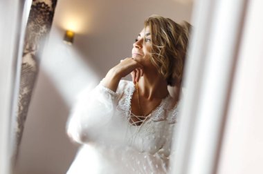 Zarif beyaz elbiseli gelin aynaya bakıyor. Gelin gülümser ve aynanın önünde poz verir. Profesyonel makyajlı ve saç stili olan güzel bir kadın. Gelin hazırlıklarının sabahı. 