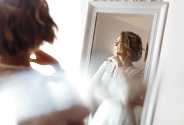 Zarif beyaz elbiseli gelin aynaya bakıyor. Gelin gülümser ve aynanın önünde poz verir. Profesyonel makyajlı ve saç stili olan güzel bir kadın. Gelin hazırlıklarının sabahı. 