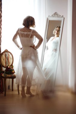 Zarif beyaz elbiseli gelin aynaya bakıyor. Gelin gülümser ve aynanın önünde poz verir. Profesyonel makyajlı ve saç stili olan güzel bir kadın. Gelin hazırlıklarının sabahı. 