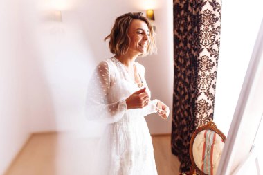 Zarif beyaz elbiseli gelin gülümser ve aynanın önünde poz verir. Güzel gelin eğleniyor ve düğününe hazırlanıyor. Makyajlı ve saç stili olan güzel bir kadın. Gelinin sabahı.