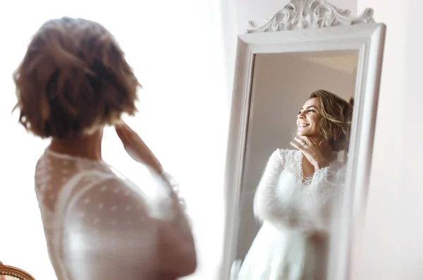Bride in elegant white dress looking in the mirror. The bride smiles and poses in front of the mirror. Beautiful woman with professional make up and hair style. Morning of the bride preparations.