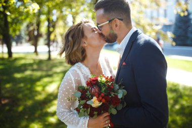 Genç çift parkta yürürken romantik anların tadını çıkarıyor. Gelin ve damat düğün günlerinde parkta poz verip öpüşüyorlar. Güzel beyaz elbiseli zarif gelin, takım elbiseli damat..