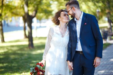 Genç çift parkta yürürken romantik anların tadını çıkarıyor. Gelin ve damat düğün günlerinde parkta poz verip öpüşüyorlar. Güzel beyaz elbiseli zarif gelin, takım elbiseli damat..