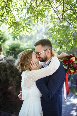 Genç çift parkta yürürken romantik anların tadını çıkarıyor. Gelin ve damat düğün günlerinde parkta poz verip öpüşüyorlar. Güzel beyaz elbiseli zarif gelin, takım elbiseli damat..