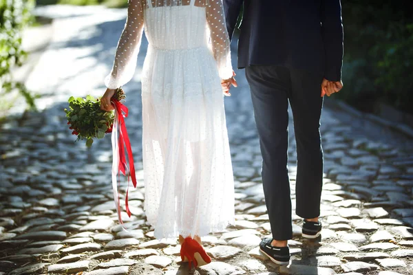 The feet of the bride and the groom with wedding bouquet. Bride in an elegant white dress and red shoes holds a stylish wedding bouquet with red and burgundy colors in her hands. Wedding details.
