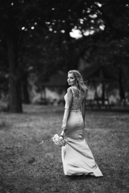 Parkta yürüyüş için bir buketi olan güzel gelinin siyah beyaz fotografı. Mutlu yeni evli kadın bahçedeki yeşil çalıların arasında duruyor. Gülümseyen gelin. Düğün günü. Moda Gelini.