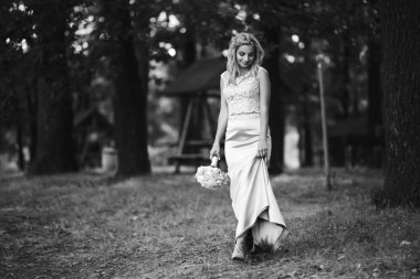Parkta yürüyüş için bir buketi olan güzel gelinin siyah beyaz fotografı. Mutlu yeni evli kadın bahçedeki yeşil çalıların arasında duruyor. Gülümseyen gelin. Düğün günü. Moda Gelini.