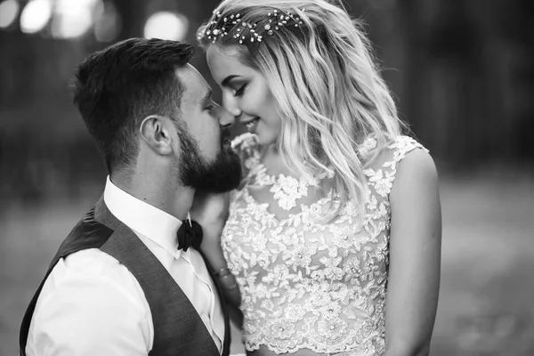 Sensual Black and white foto of bride and groom. Stylish couple of happy newlyweds posing in the park on their wedding day. Handsome bearded groom admires and kisses pretty bride. Together.