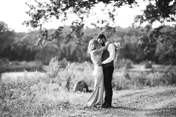 Sensual Black and white foto of bride and groom. Stylish couple of happy newlyweds posing in the park on their wedding day. Handsome bearded groom admires and kisses pretty bride. Together.