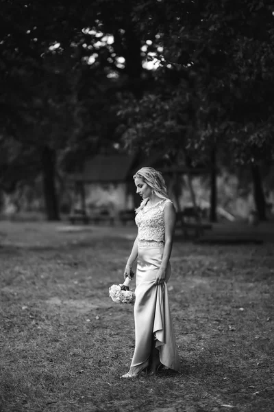Black and white foto of beautiful bride with a wedding bouquet for a walk in the park. Happy newlywed woman stands among green bushes in the garden. Smiling bride. Wedding day. Fashion bride.