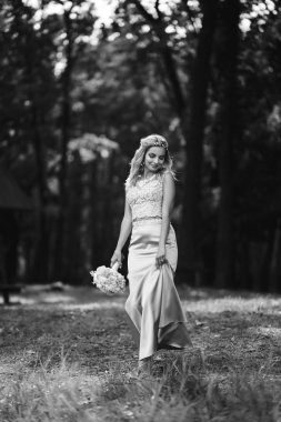 Parkta yürüyüş için bir buketi olan güzel gelinin siyah beyaz fotografı. Mutlu yeni evli kadın bahçedeki yeşil çalıların arasında duruyor. Gülümseyen gelin. Düğün günü. Moda Gelini.