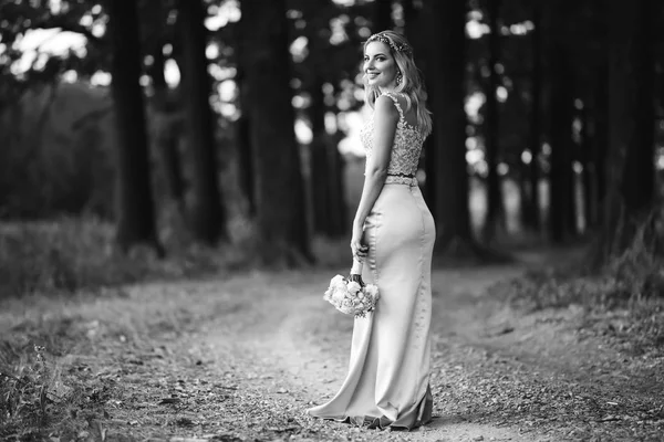 Black and white foto of beautiful bride with a wedding bouquet for a walk in the park. Happy newlywed woman stands among green bushes in the garden. Smiling bride. Wedding day. Fashion bride.