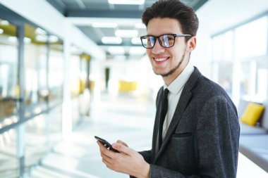 Elinde telefonla kameraya bakıp gülümseyen bir iş adamı. Modern ofiste elinde telefonla duran takım elbiseli şık bir adam. Gözlüklü genç ofis çalışanı cep telefonu kullanıyor..