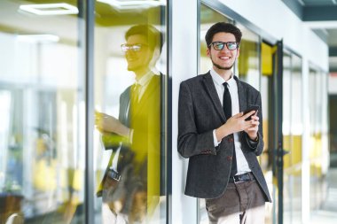 Elinde telefonla kameraya bakıp gülümseyen bir iş adamı. Modern ofiste elinde telefonla duran takım elbiseli şık bir adam. Gözlüklü genç ofis çalışanı cep telefonu kullanıyor..