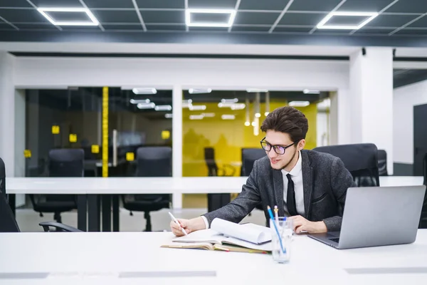 Young office manager working with papers and computer working at office. Businessman writes, reviews and analyze documents, plans. Stylish man in a suit working with new startup project.